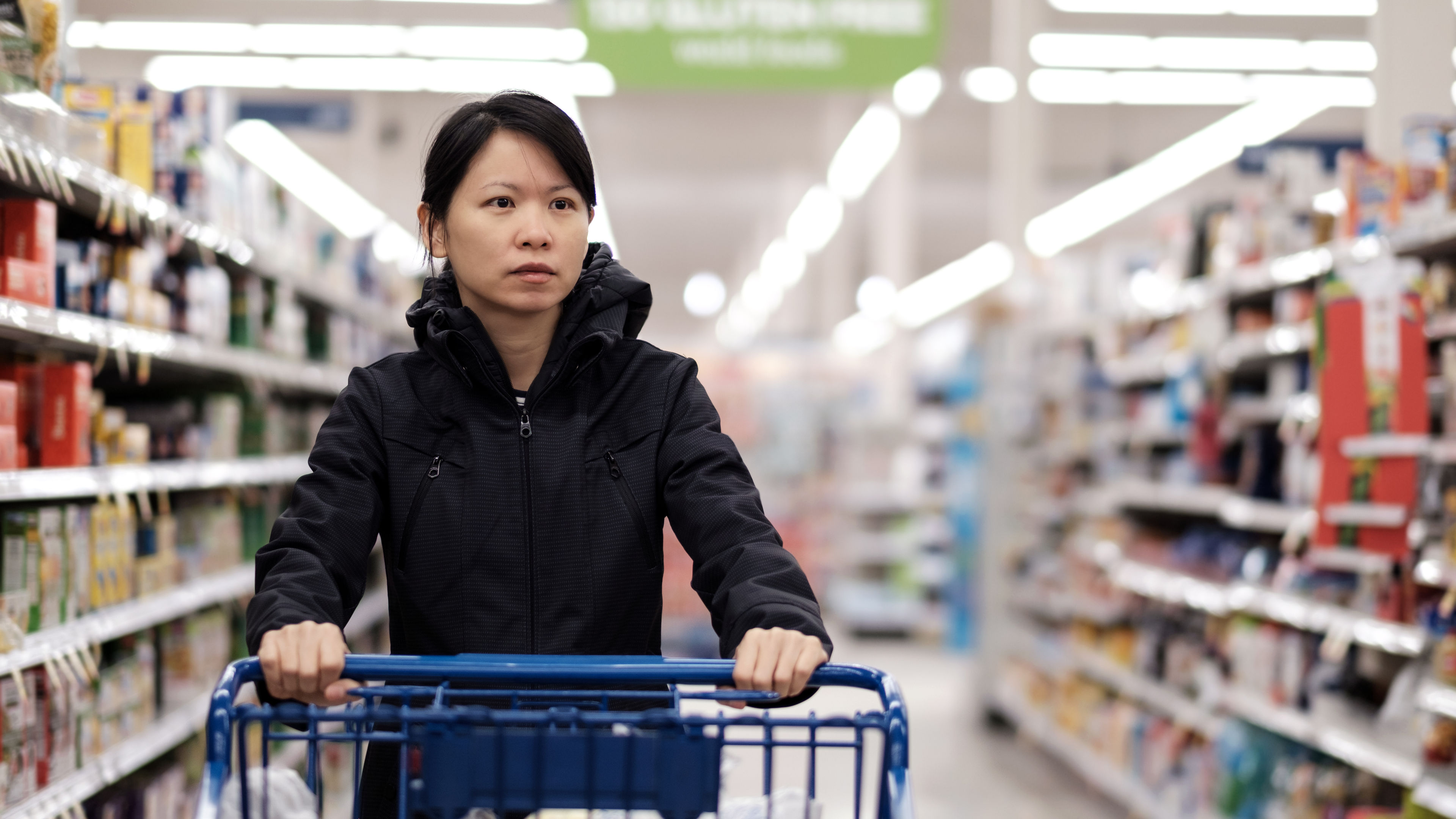 A woman inside a supermarket, pushing a trolley bag filled with various products.