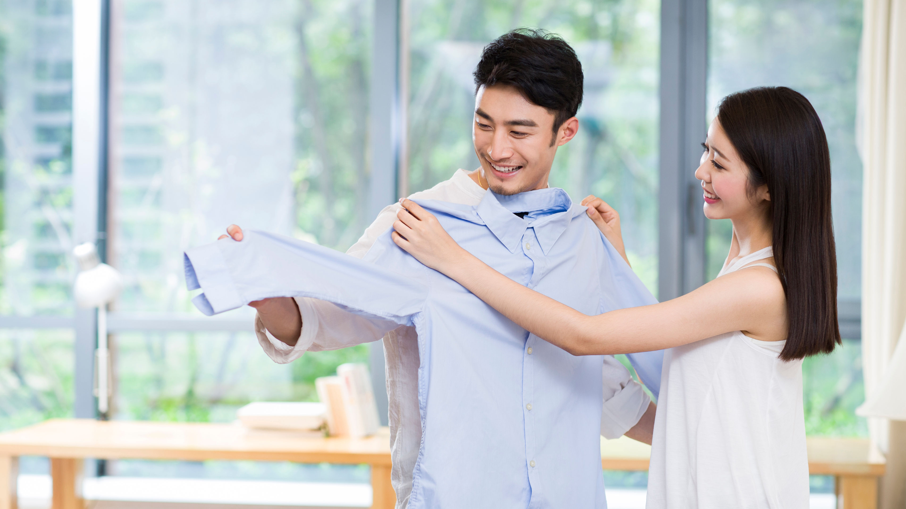 A cheerful couple stands in their cozy living room, holding up a light blue shirt together 