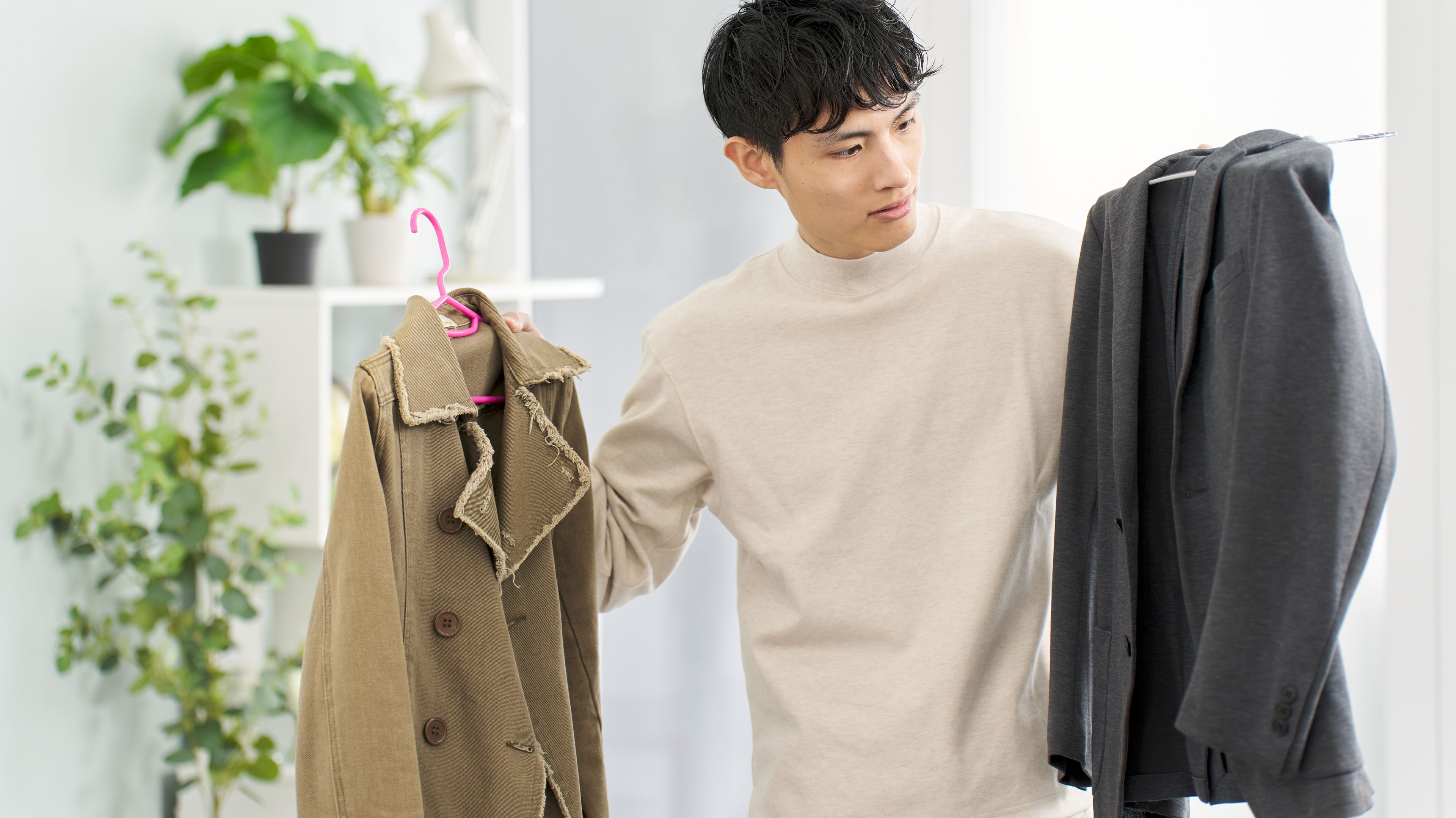 A young man stands in a well-lit room holding two coats on hangers 