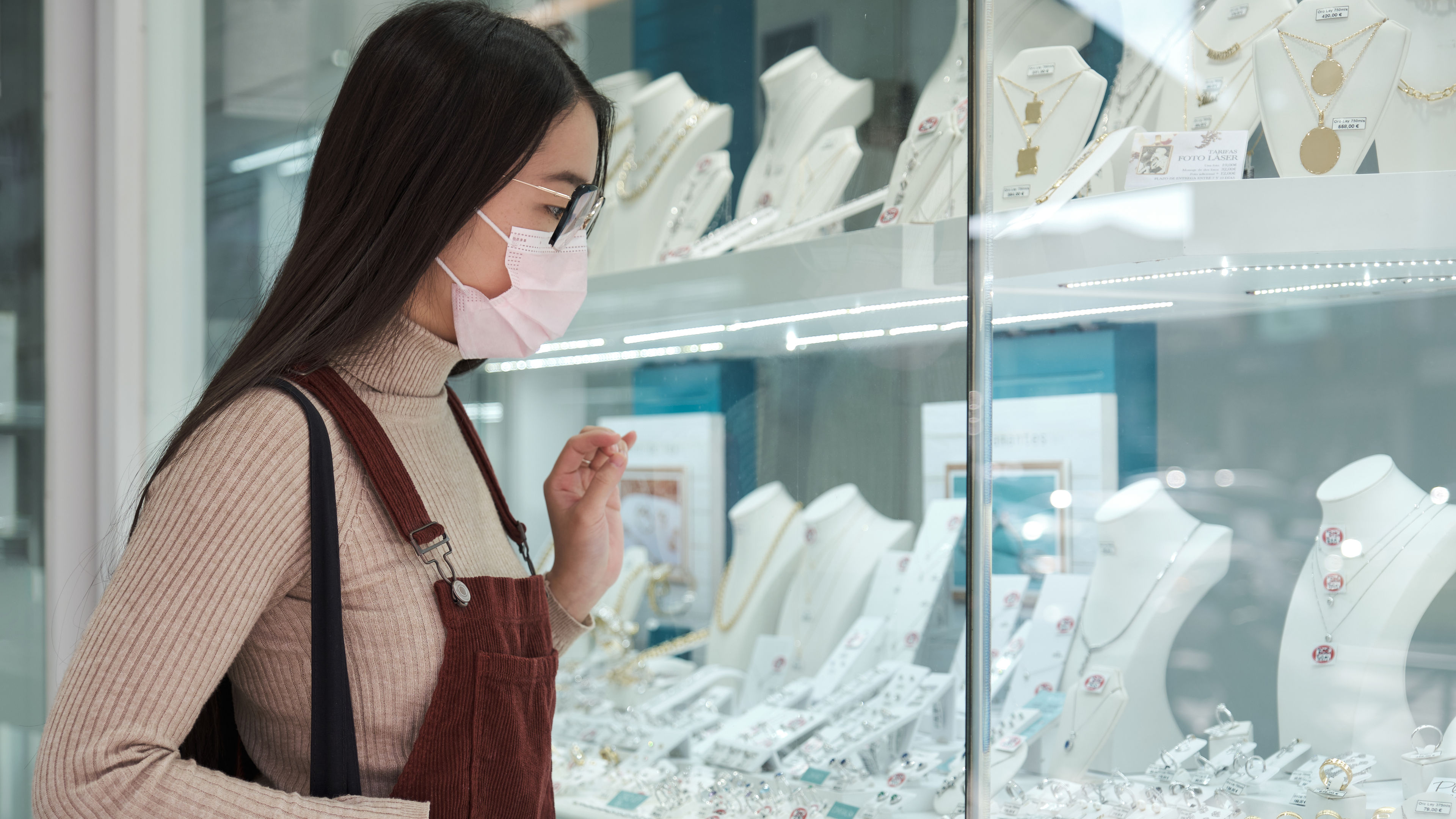 Wearing a face mask and chic outfit, the woman carefully examines sparkling necklaces and earrings 