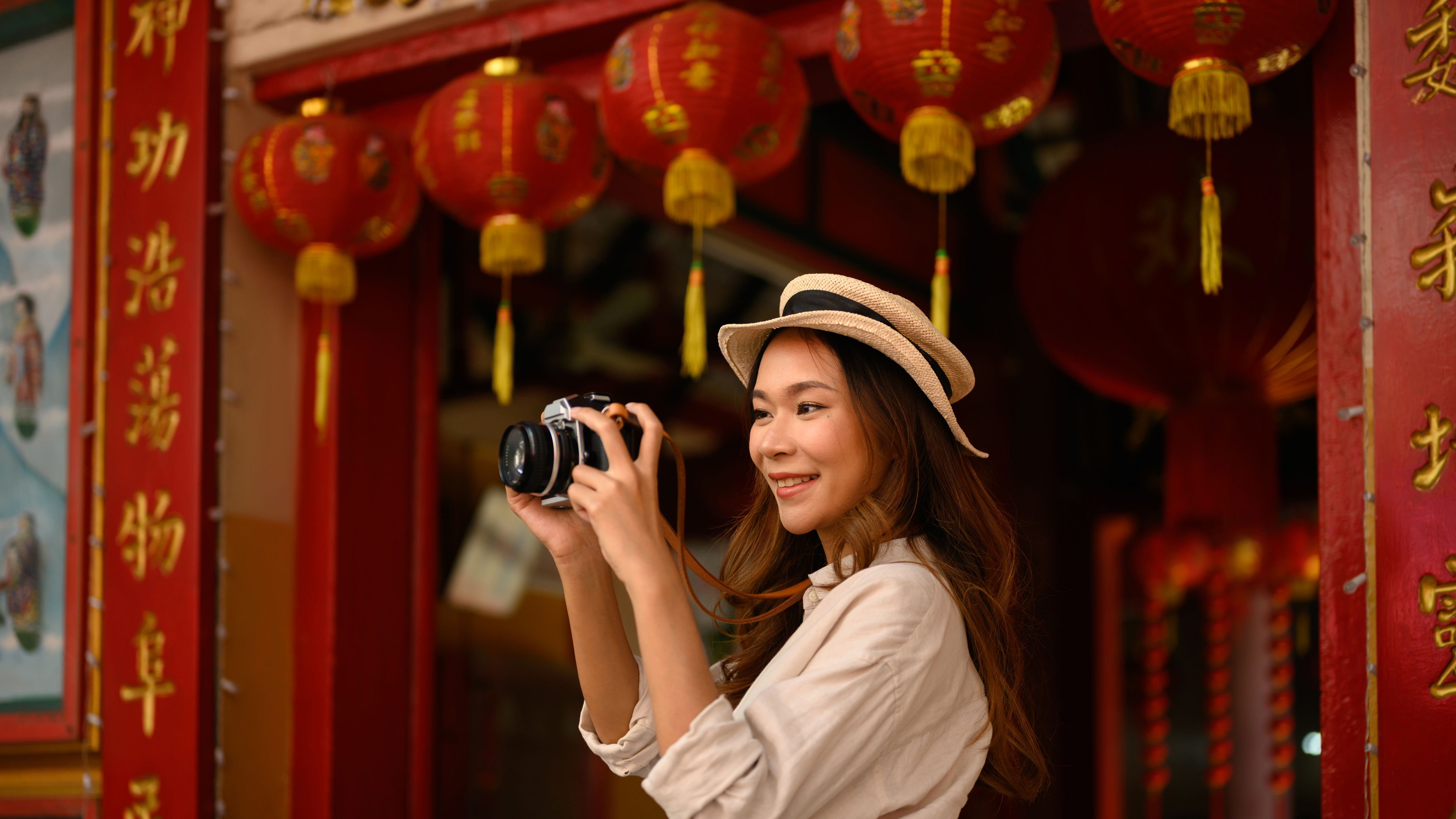 一位面帶微笑嘅年輕女子企喺中國寺廟前，上面裝飾住靚嘅紅燈籠。 

