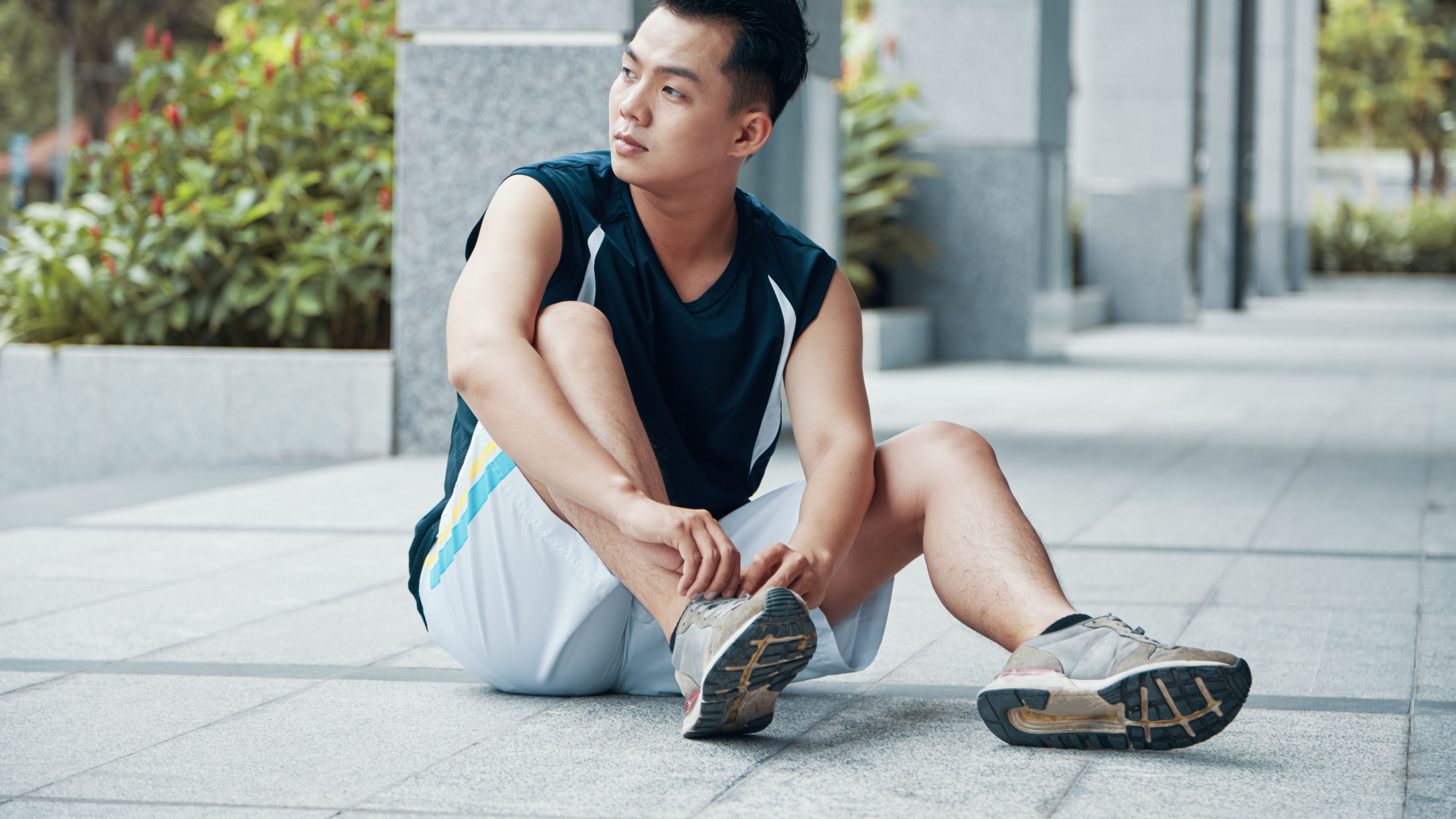 Attractive Asian guy in sportswear tying laces on sneakers and looking away while sitting on ground during outdoor training 