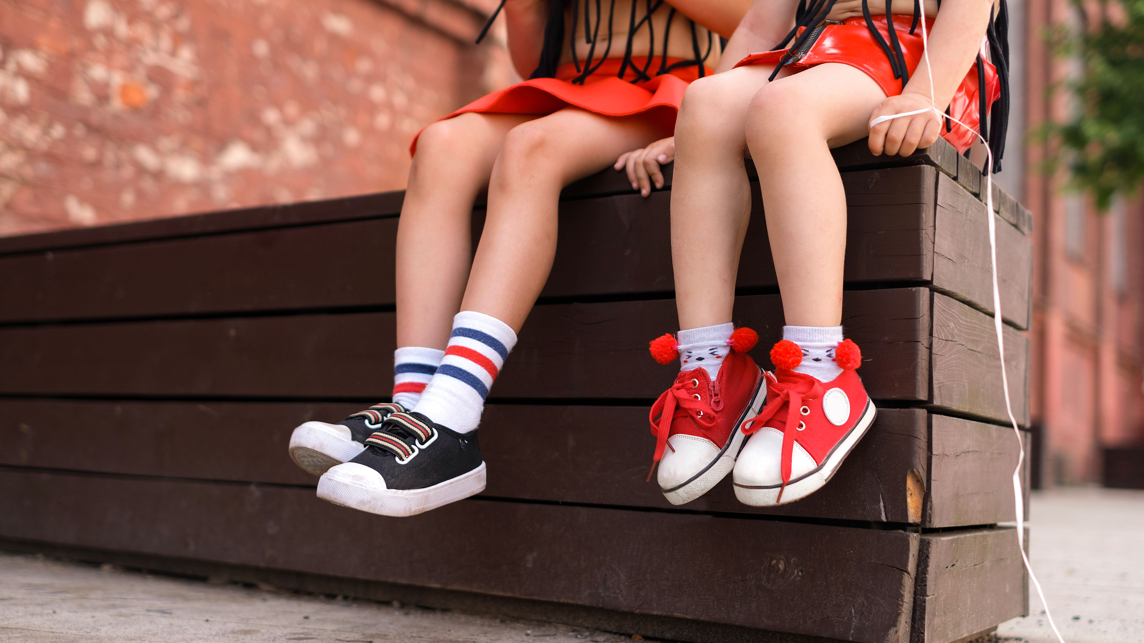 Two young girls are sitting on a wooden bench wearing colorful sneakers 
