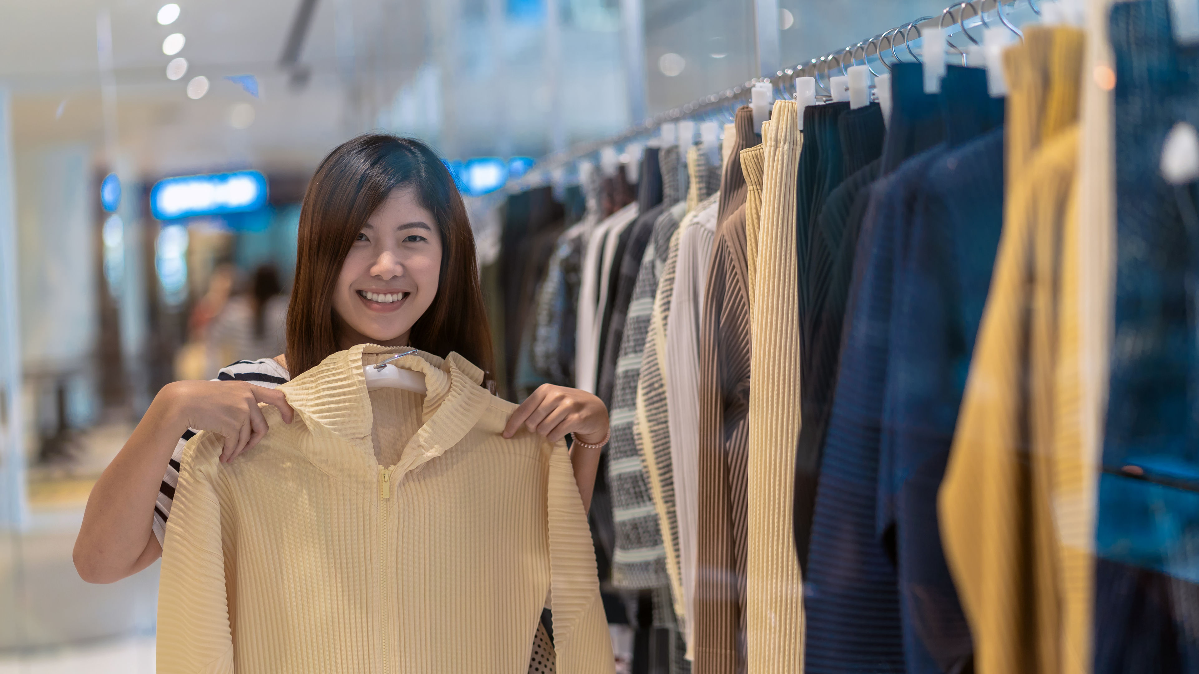 A smiling shopper holds up a light yellow sweater from a store rack 
