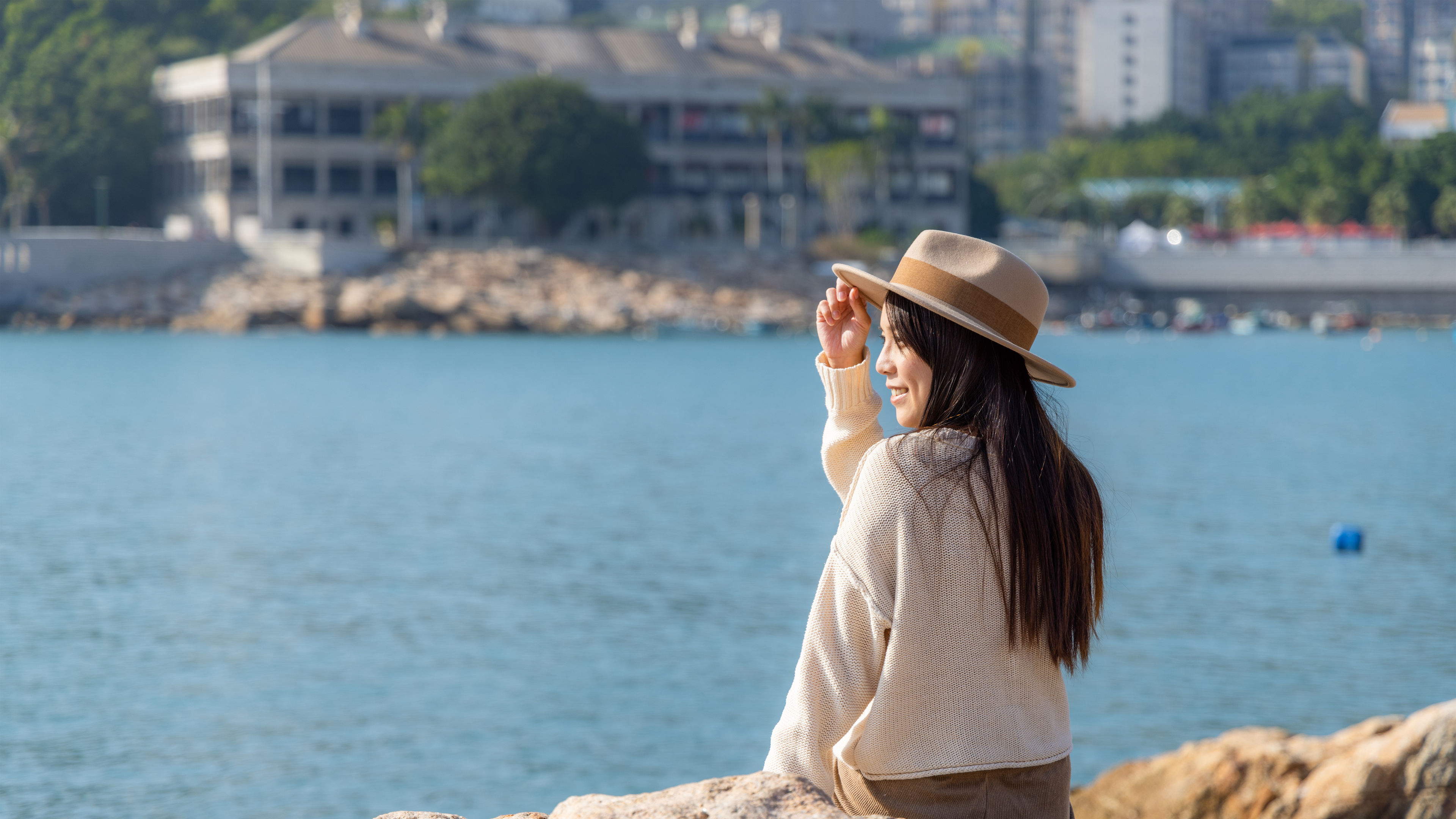 Sitting on rocky terrain, a woman in a cozy sweater and hat gazes at the tranquil sea 